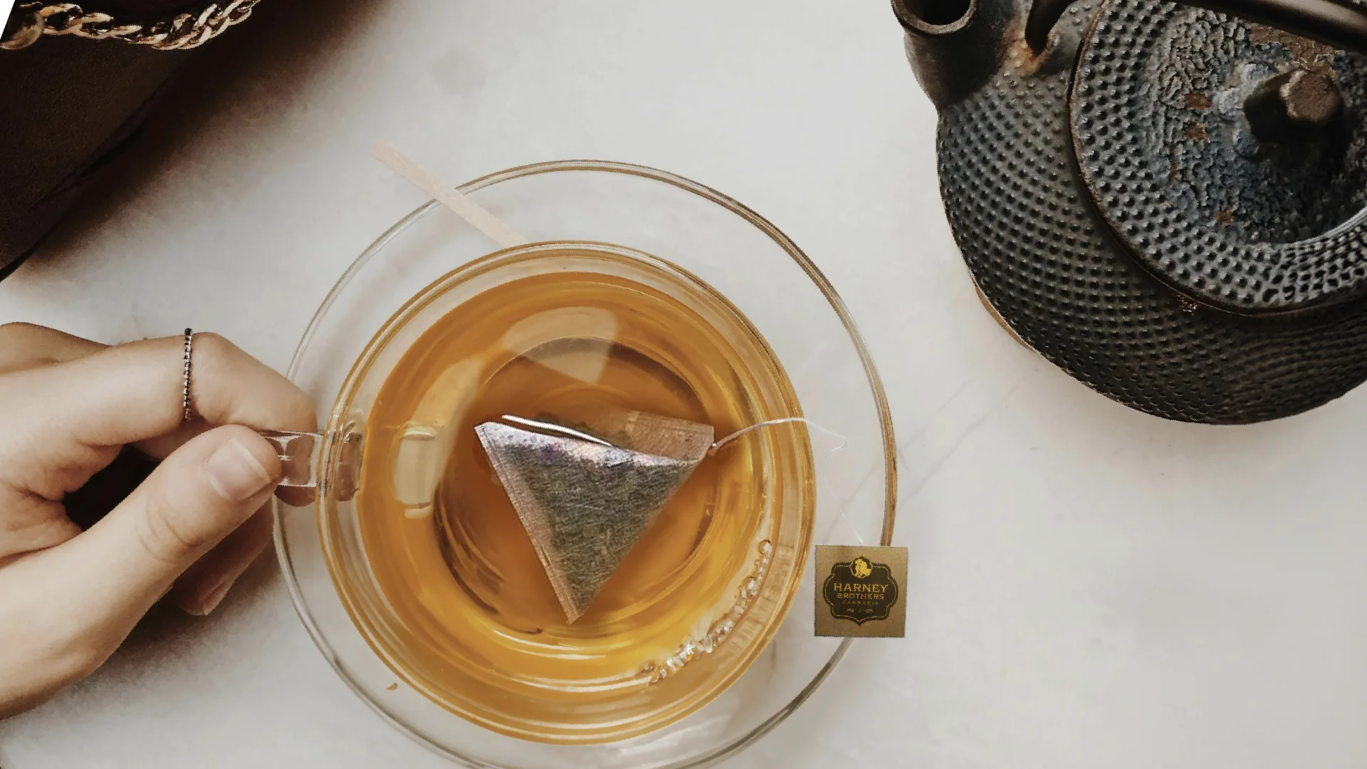 A glass cup of tea with a Harney Brothers tea sachet steeping beside a black iron teapot.