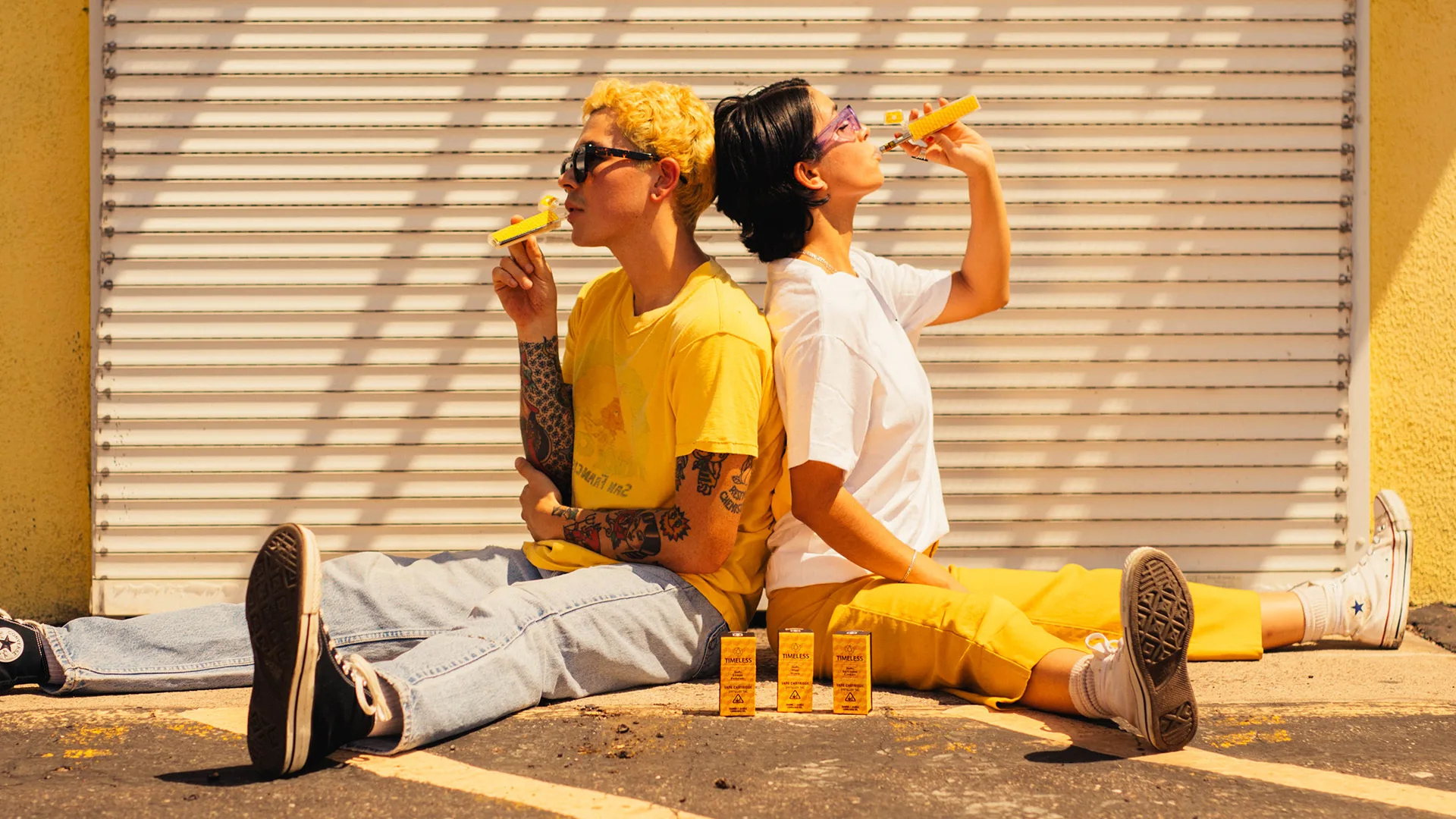 Two women sitting, both dressed in yellow, smoking vape carts from Timeless.
